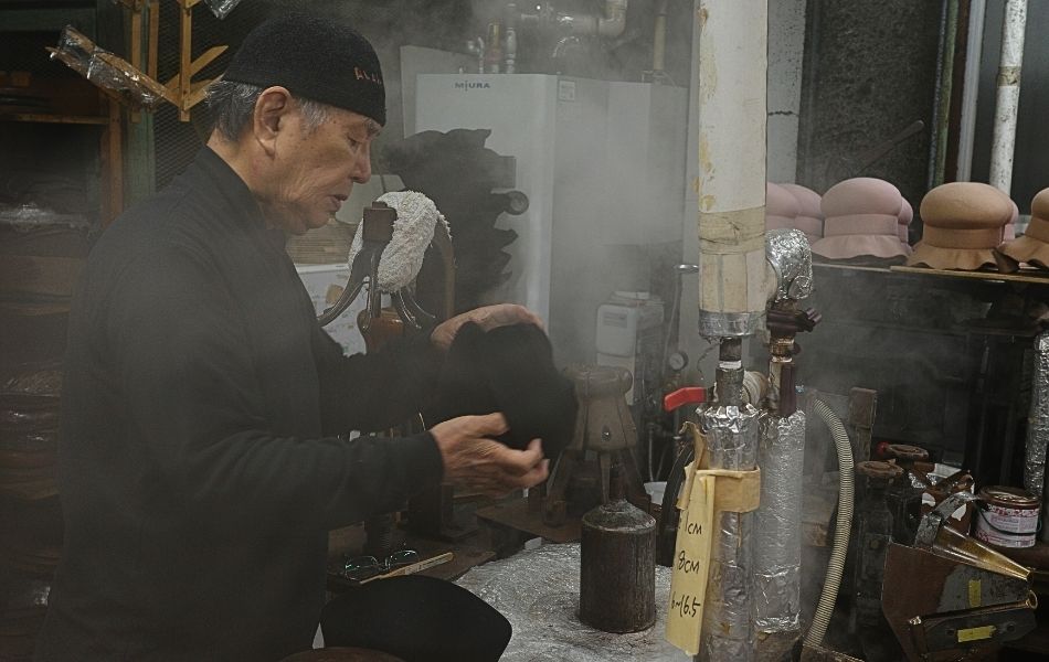 蒸気の立ちこめる工房で帽子の形を確かめる森安会長。木型製法による手仕事の現場。